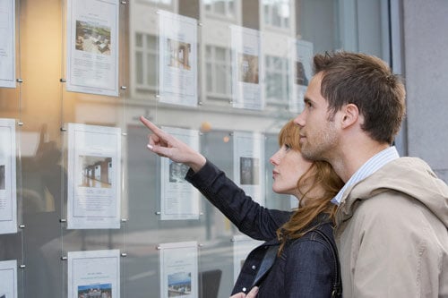 Man standing closely behind woman while they both look at pictures of properties outside an estate agent window